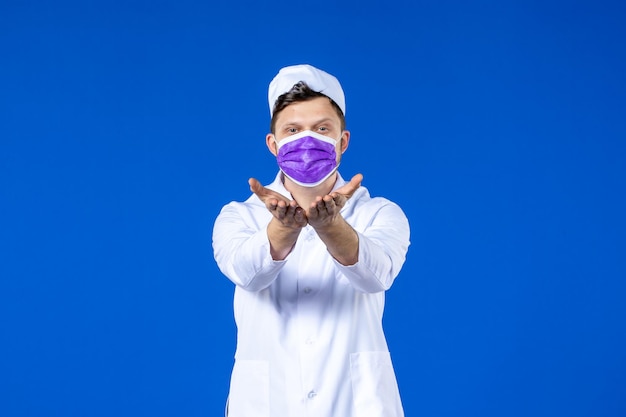 Вид спереди на мужчину-врача в медицинском костюме и фиолетовой маске, отправляющего поцелуи на синем