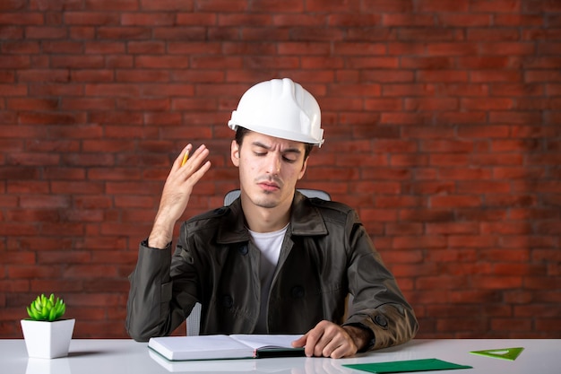 Front view male engineer sitting behind his working place in white helmet document plan business corporate job builders contractor property agenda