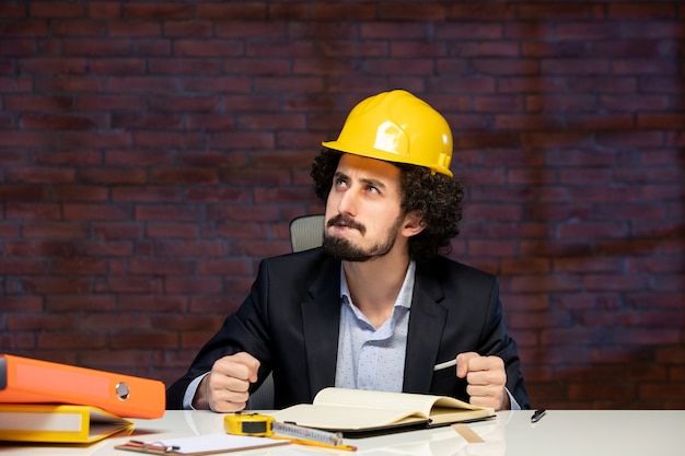 Vista frontale dell'ingegnere maschio seduto dietro il posto di lavoro in tuta e casco giallo costruttore di piani appaltatore aziendale occupazione lavoro progetto aziendale lavoro