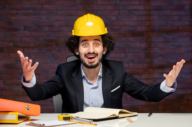 вид спереди мужчины-инженера, сидящего за рабочим местом в костюме и желтом шлеме, план бизнес-строитель, корпоративный проект работы подрядчика