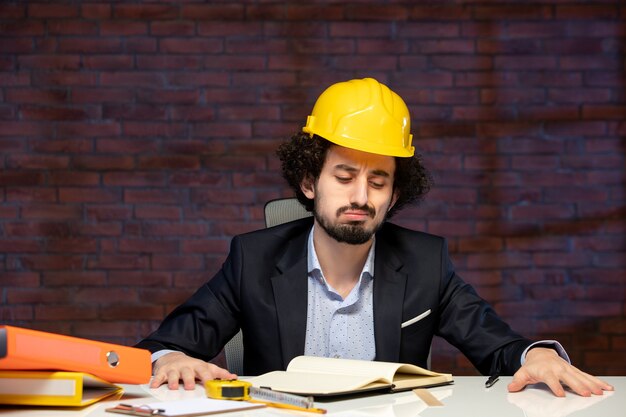 Vista frontale dell'ingegnere maschio seduto dietro il posto di lavoro in tuta e casco giallo piano progetto aziendale lavoro aziendale appaltatore costruttore di occupazione