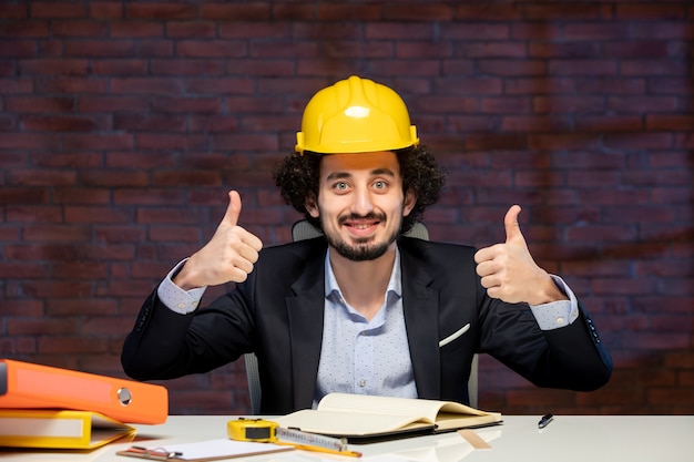 front view of male engineer sitting behind working place in suit and yellow helmet plan business project corporate job occupation builder work