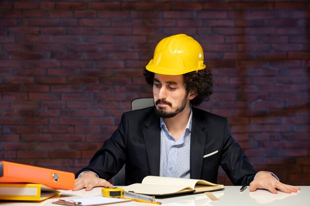 Vista frontale dell'ingegnere maschio seduto dietro il posto di lavoro in tuta e casco giallo piano progetto aziendale lavoro appaltatore occupazione costruttore lavoro aziendale