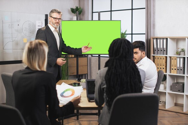 Photo front view of mature confident bearded businessman showing statistic datas on green screen