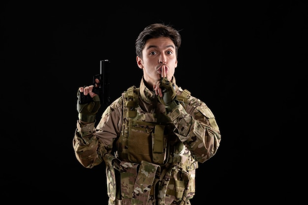 Front view of military serviceman in uniform with gun black wall