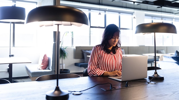 Front view of a mixed race businesswoman working in a modern office, sitting by a desk and using a laptop computer. Social distancing and self isolation in quarantine lockdown