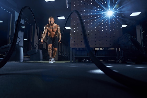 Front view of muscular brunette shirtless man doing battle rope training in gym in dark atmosphere.