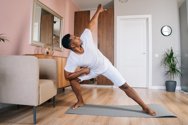 Front view of muscular strong African-American man doing side lunge exercise at home during working out standing on yoga mat at domestic room, looking away. Concept of sport training at home gym.