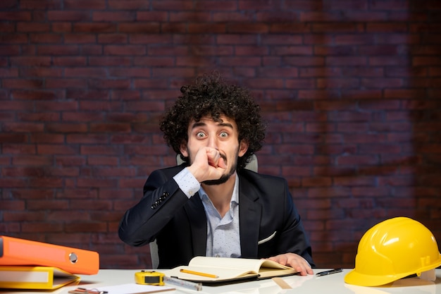 Foto vista frontale dell'ingegnere maschio nervoso seduto dietro il suo posto di lavoro in tuta appaltatore del piano lavoro progetto aziendale lavoro occupazione costruttore business