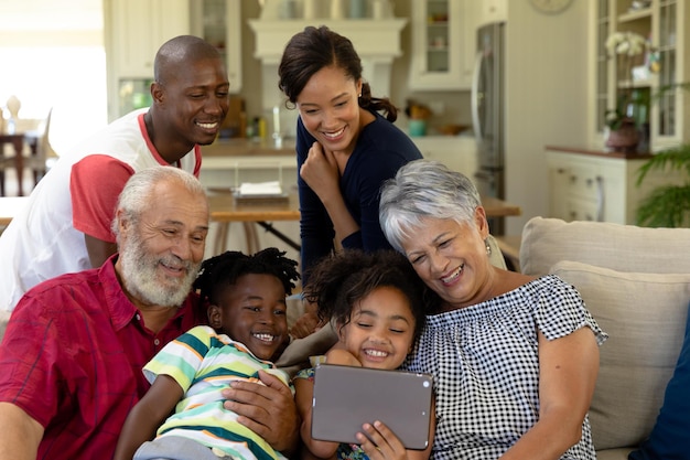 写真 自宅のリビングルームでソファに集まり、孫娘が持っているタブレットコンピューターを見ている多世代の混血家族の正面図