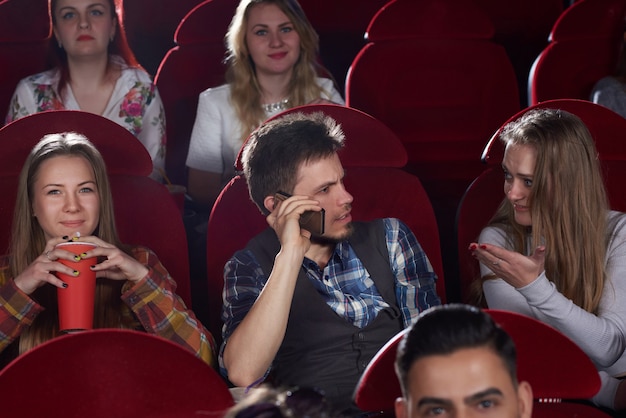 Фото Вид спереди группы людей, смотрящих фильм в кинозале, брюнет разговаривает по телефону, сердитая и шокированная женщина жестикулирует, останавливая его. досуг и культура юных девушек и юношей.