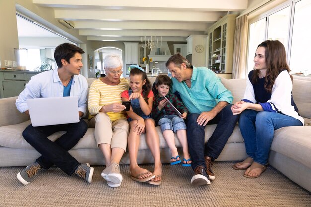 Фото Вид спереди на шесть членов кавказской семьи из нескольких поколений, сидящих дома на диване в гостиной и использующих ноутбук, смартфон и планшетный компьютер. семья наслаждается временем дома, жизнью