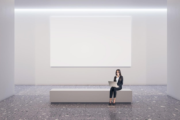 Фото Вид спереди на пустой белый плакат с местом для вашего логотипа на светлом фоне стены в просторном зале галереи с деловой женщиной, использующей ноутбук и сидящей на каменной скамейке на бетонном полу, макет