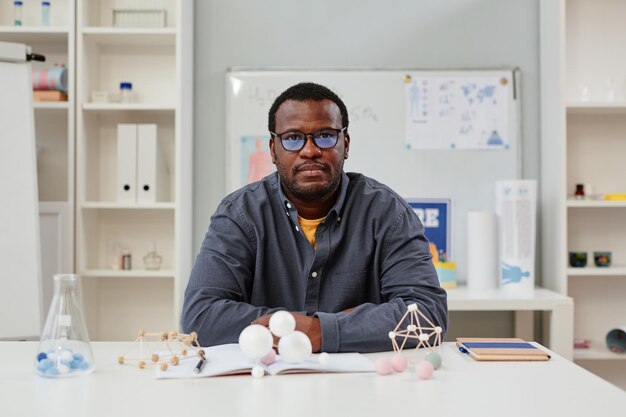 Photo front view portrait of african american teacher sitting at desk in chemistry class with molecule models copy space