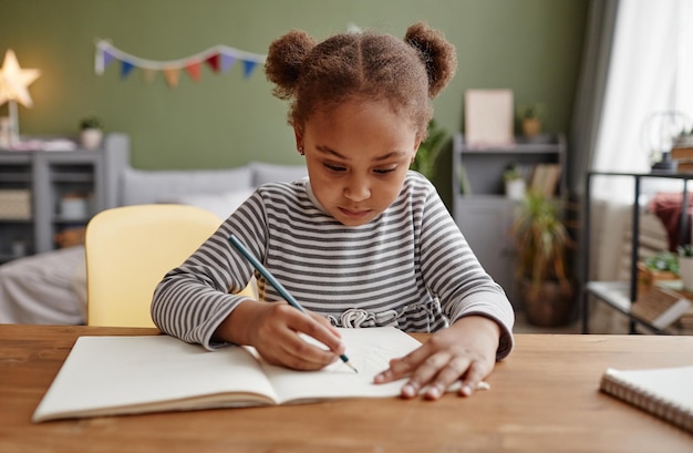 Photo front view portrait of cute african-american girl doing homework while sitting at wooden desk, copy space