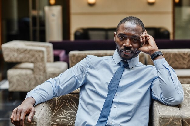 Front view portrait of handsome African-American businessman looking at camera while relaxing at luxurious hotel lobby, copy space