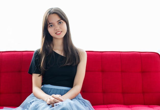 Front view portrait shot of an attractive smiling junior young Thai-Turkish teenager wearing trendy casual clothes sitting on the red couch and looking at camera in studio with windows background.