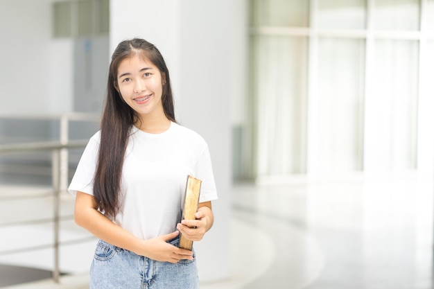 캠퍼스 건물에 책을 들고 등교하는 편안한 캐주얼 복장을 한 젊고 쾌활한 아시아 청소년 여성 대학생의 전면 초상화