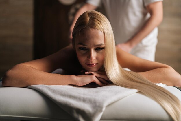 Front view of relaxed blonde young woman lying down on massage table during back massage at spa