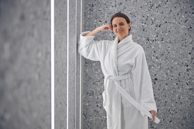 Photo front view of a relaxed woman in a bathrobe leaning her elbow against the wall