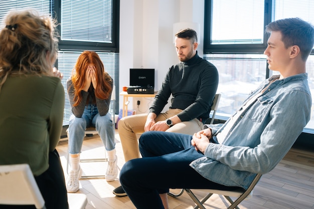 Front view of sad frustrated young woman sharing problem during group therapy sitting with head bowed. Concept of group consulting of mental health problem with psychologist.