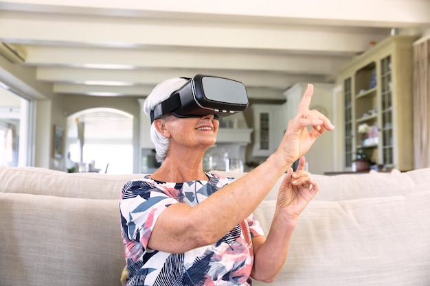 Foto vista frontale di una donna caucasica anziana con un visore vr, che tocca uno schermo virtuale con il dito, seduta su un divano in soggiorno, sorridente. famiglia che si gode il tempo a casa, concetto di stile di vita