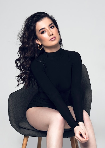 Front view of a sensual young woman with hairstyle wear black bodysuit sitting on chair, looking at camera, on a grey background. Vertical view.