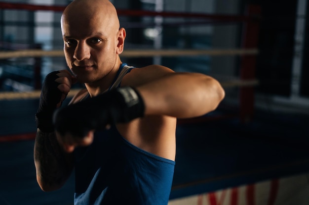 Front view of serious aggressive boxer male wearing bandages punching air to camera in sport club with dark interior, on background of ring. Professional fighter fighting shadow looking at camera.