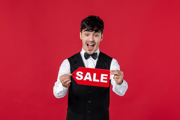 Front view of shappy guy waiter in a uniform with butterfly on neck holding sale icon on isolated red wall