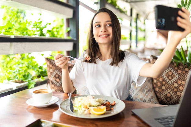 Front view of smiling attractive woman eating tasty food in cafe and doing selfie on modern phone.