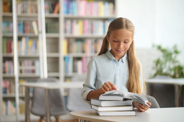 Photo front view of a smiling joyous young girl perusing a textbook at the desk at a public library