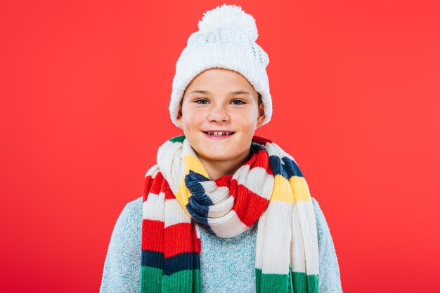 Front view of smiling kid in hat and scarf isolated on red