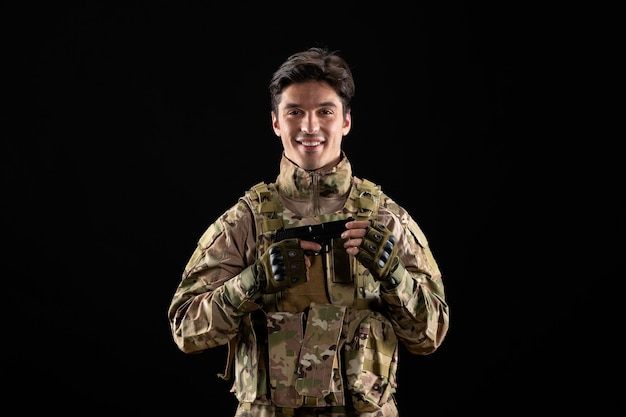 Front view of smiling military serviceman in uniform with gun black wall