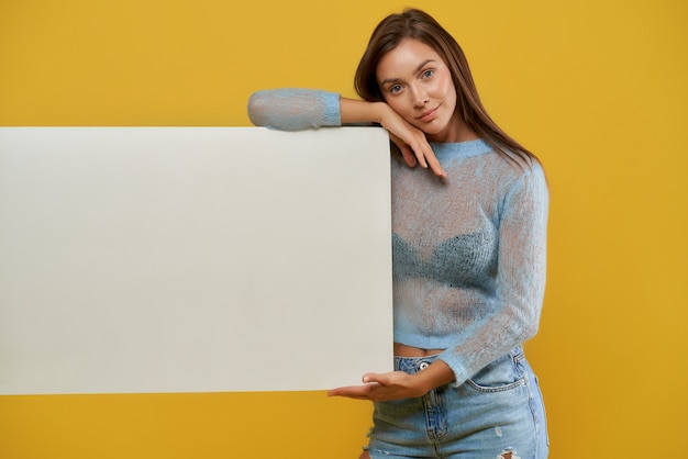 Front view of smiling pretty lady demonstrating cardboard blank poster. Charming young woman land resting head on hand. Isolated on yellow  wall. Concept of advertising.