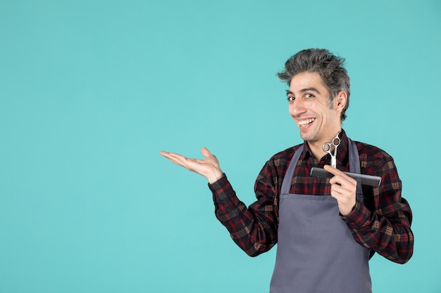 Front view of smiling young barber wearing gray apron and holding comb and scissor pointing something on the right side on blue color background