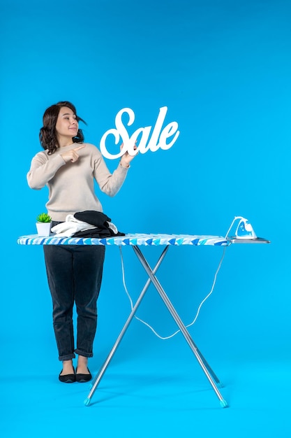 Front view of smiling young woman standing behind the ironing board and showing sale icon on blue wall