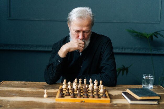 Front view of thoughtful bearded mature pensioner male thinking about chess move sitting at home and playing chess alone