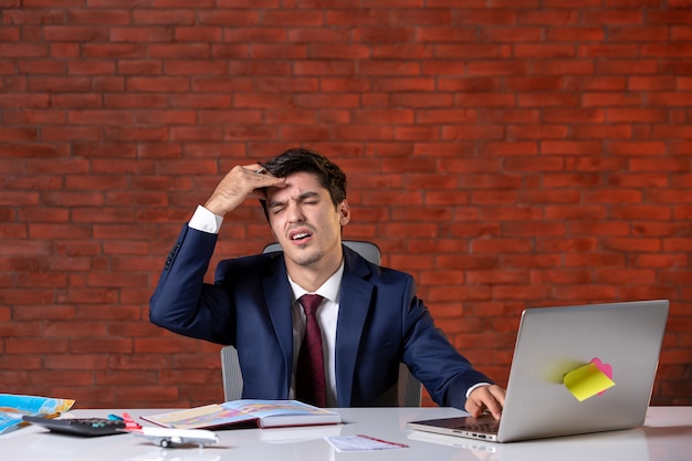 front view of tired travel agent sitting behind his working place in suit and using laptop corporate occupation service tourism global agency project