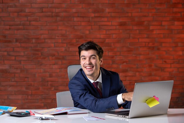 front view of travel agent sitting behind his working place in suit and using laptop corporate occupation tourism project service global agency assistant