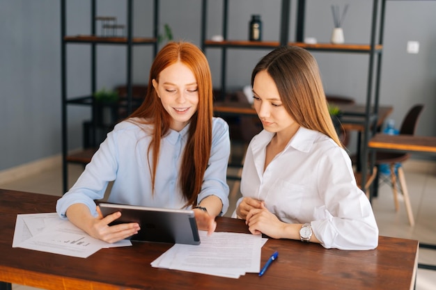 Front view of two friendly young business women working using digital tablet at meeting desk with job documents at office