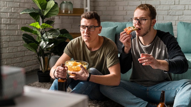 Photo front view of two male friends having beer with snacks and watching sports on tv