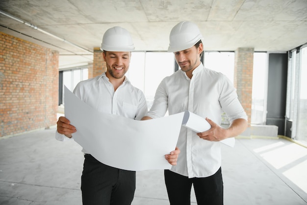 A front view of two smart architects with white helmets reviewing blueprints at a construction site on a bright sunny day