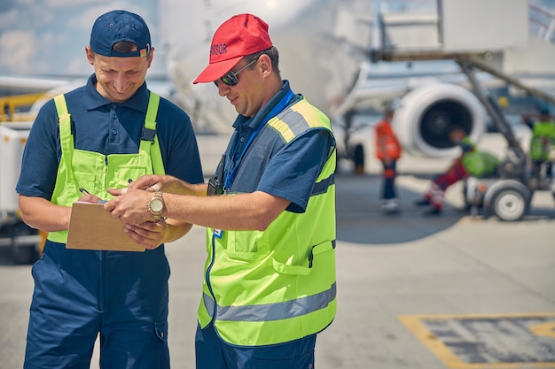 Front view of two smiling airport male workers filling out a form on a clipboard