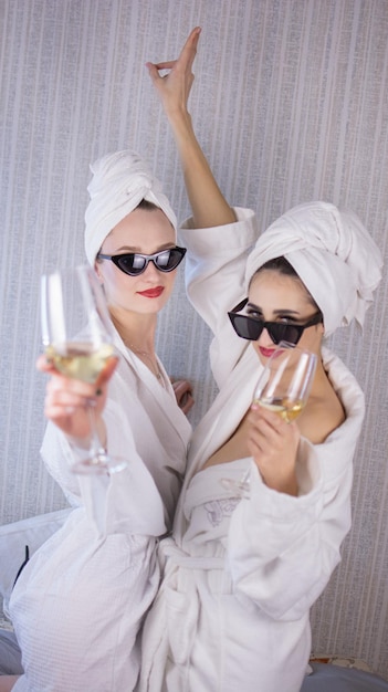 Photo front view of two smiling female friends with towels and in bathrobes sitting on bed and doing