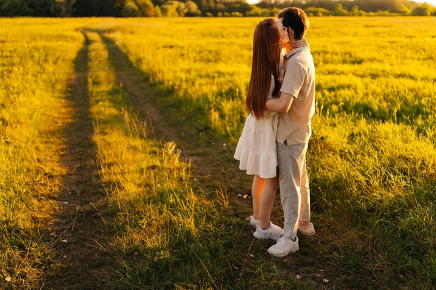 Front view of unrecognizable happy young couple in love standing hugging on beautiful green meadow in summer evening during golden sunset with soft sunlight