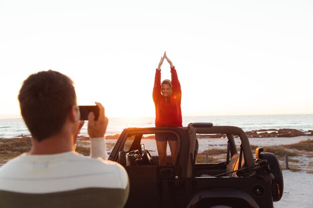 Front view van een blank echtpaar buiten een open auto, met zonsondergang op het strand op de achtergrond, de man die een foto maakt van zijn partner die handen opheft en glimlacht