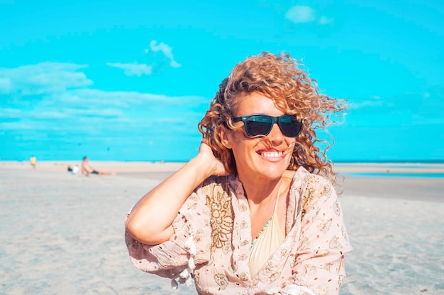 Front view van een mooie vrouw die op het strand zit Blonde vrouw met krullend haar die in de wind waait ontspant zich kijkt en glimlacht