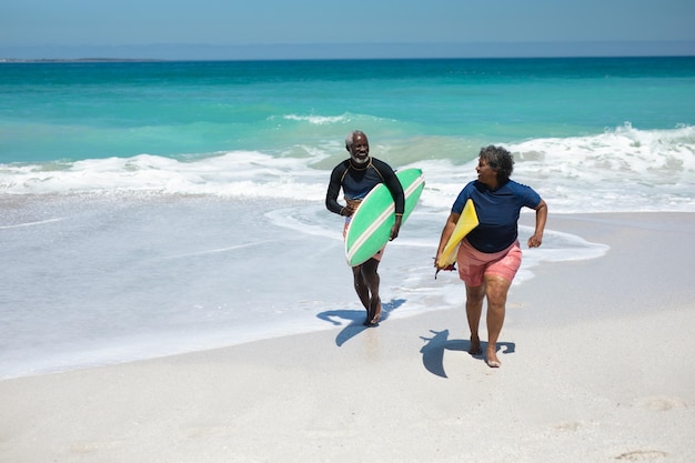 Front view van een oudere Afro-Amerikaanse echtpaar op een strand in de zon, met surfplanken onder hun armen en lopen, naar elkaar kijken en glimlachen, met blauwe lucht en rustige zee op de achtergrond