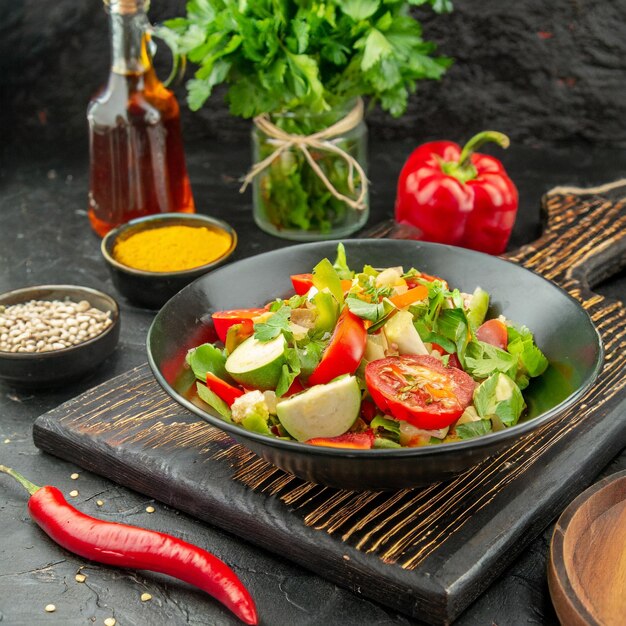 Photo front view of vegan salad with fresh ingredients in a plate and pepper on black cutting board