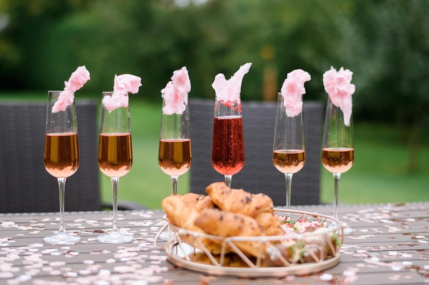 屋外での結婚式の朝食時に焼きクロワッサンのプレートの近くのテーブルに置かれたピンクの花で飾られたカラフルなバブルシャンパンとワイングラスの正面図
